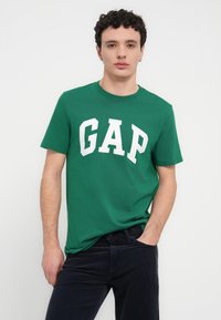 Πράσινο βαμβακερό μπλουζάκι με λευκό λογότυπο "GAP" τυπωμένο εμφανώς στο μπροστινό μέρος. Κοντά μανίκια και σχέδιο με στρογγυλή λαιμόκοψη.
