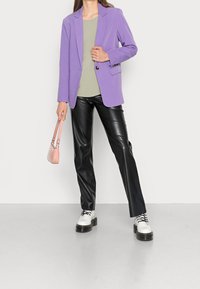 Blazer lavanda, top verde chiaro, pantaloni in ecopelle nera, scarpe platform bianche e una piccola borsa rosa. Texture lisce, vestibilità su misura.