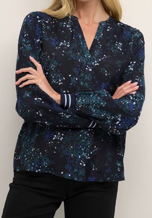 Femme portant un chemisier à manches longues à motifs floraux foncés avec des poignets rayés, les bras croisés, et un pantalon noir sur un fond uni.