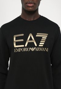 Svart hettegenser laget av bomull, med et stort gulllogo "EA7 EMPORIO ARMANI" på forsiden. Ribbestrikket halslinning og mansjetter.