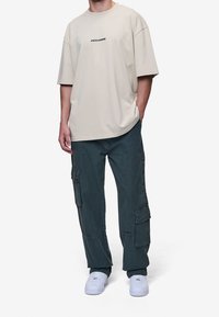 Beige oversized tričko s černým logem "PEGADOR", spárované s tmavě zelenými cargo kalhotami s několika kapsami a bílými teniskami.