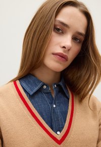 Suéter beige con ribete rojo en forma de V sobre una camisa de mezclilla. El suéter es de punto, mientras que la camisa tiene un cuello abotonado y costuras en contraste.