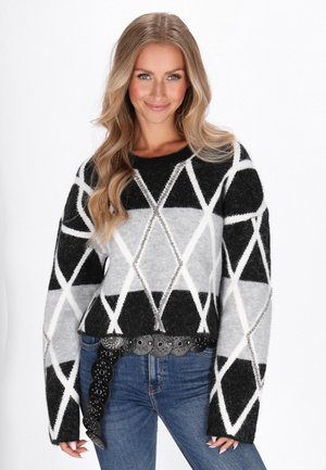 Kvinde med langt blondt hår iført en sort, grå og hvid sweater med geometrisk mønster og blå jeans med sort naglet bælte.