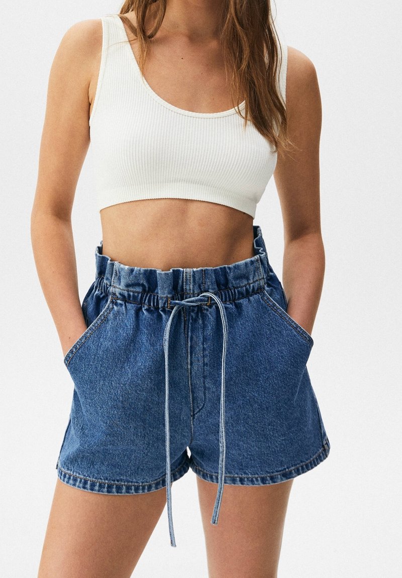 Femme portant un haut court côtelé blanc et un short en jean bleu taille haute avec une taille à cordon et les mains dans les poches.