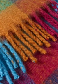 Multikrāsaina vilnas sega, kurā ir oranži, zili un sarkani toņi ar ķemmētu malu, izceļot mīkstu, teksturētu virsmu.