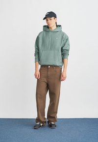 Πράσινη βαμβακερή φόρμα με τσέπη καγκουρό, συνδυασμένη με καφέ τζιν παντελόνι. Μαύρο καπέλο και παπούτσια ολοκληρώνουν την εμφάνιση. Απλός, καθημερινός σχεδιασμός.