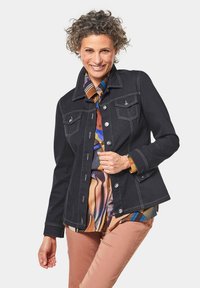 Dunkelblaue Jeansjacke mit Druckknöpfen, zwei Brusttaschen und kontrastierendem weißen Nähten, über einem bunten gemusterten Hemd getragen.