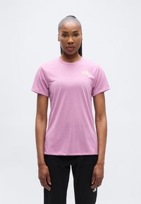 Γυναίκα με σκούρο δέρμα και ίσια μαλλι ά σε αλογοουρά, που φοράει ανοιχτό μοβ μπλουζάκι The North Face και μαύρο παντελόνι, μπροστά σε απλό φόντο.