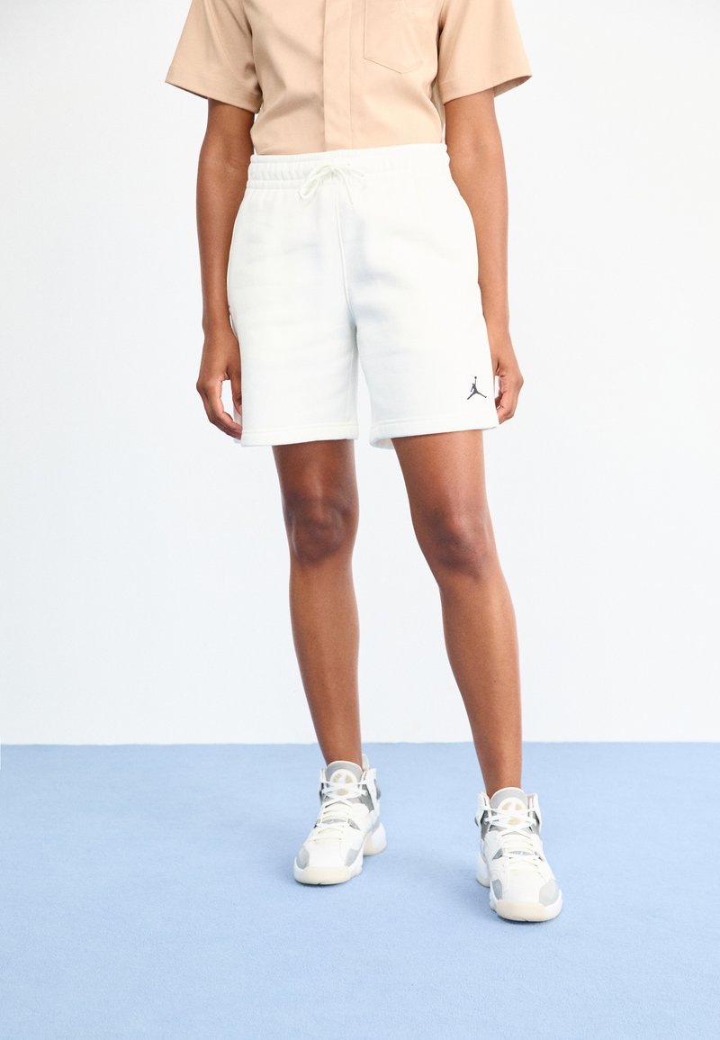 Persoon in een beige shirt, witte shorts met touwtjes en het Jordan-logo, en witte hoge sneakers staat op een lichtblauwe vloer tegen een witte achtergrond.