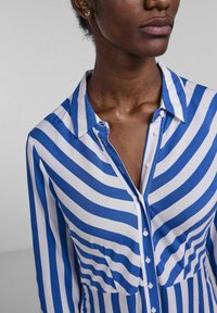 Chemise rayée bleu et blanc, avec un col, fermeture à boutons et tissu doux. Présente une silhouette ajustée et un détail de poche poitrine.