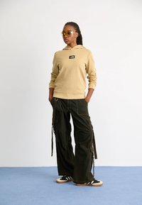 Beżowa bluza z kapturem z czarnym logo, zestawiona z ciemnymi spodniami cargo z wieloma kieszeniami i regulowanymi paskami, noszona z czarnymi butami.