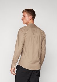 Camicia béige a maniche lunghe realizzata in tessuto morbido, con colletto a punta e polsini abbottonati, orlo curvo e dettaglio di piega posteriore.
