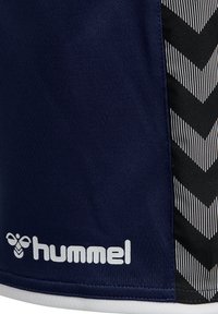 Ciemnoniebieska tkanina z białym logo „hummel” i ikoną pszczoły, obok wzoru w czarno-białe szewrony po prawej stronie.
