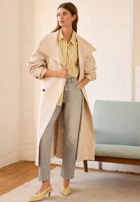 Cappotto trench beige sopra una maglietta a righe gialle e bianche, abbinato a jeans dritti grigi chiari e décolleté gialli.