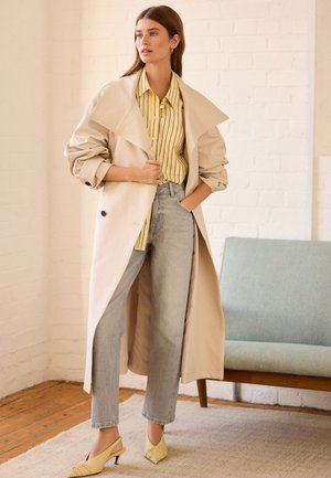 Beige trenchcoat over en gul og hvid stribet skjorte, parret med lysegrå straight-leg jeans og gule slingback hæle.