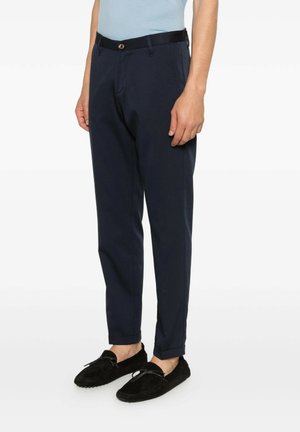 Uomo che indossa pantaloni su misura blu navy con cintura nera e mocassini in suede neri, in piedi davanti a uno sfondo bianco uniforme.