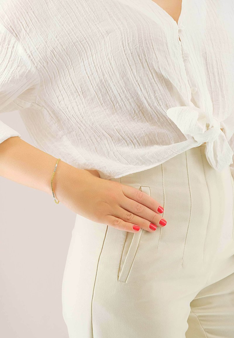 Blouse blanche froissée avec un nœud à l'avant, pantalon crème ajusté avec des poches latérales, et un bracelet doré au poignet avec des ongles rouges.