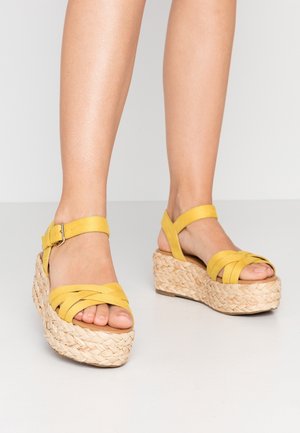 Piernas con sandalias de plataforma amarillas con tiras y suelas de paja tejida, de pie sobre un suelo blanco contra un fondo liso.