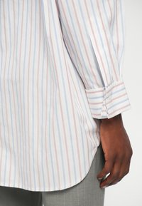 Kreklu ar garām piedurknēm, kuram ir zilas un sarkanas vertikālas svītras uz baltas fona. Tam ir kustīga apakšmala un pogājamas aproces detaļa.