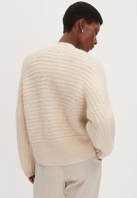 Hellbeige gestrickter Pullover mit einer strukturierten, gerippten Musterung, oversized Passform und abgesenkten Schultern. Der Stoff scheint weich und leicht flauschig zu sein.