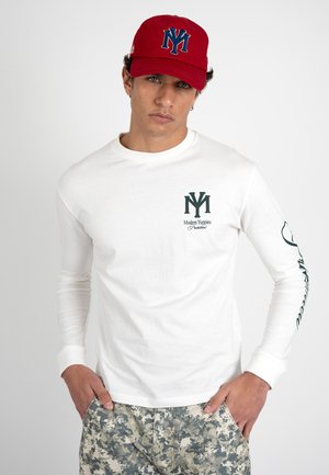 Young man wearing a red cap with blue logo, white long-sleeve shirt with black text and logo, and camo-patterned pants, standing with hands in pockets.