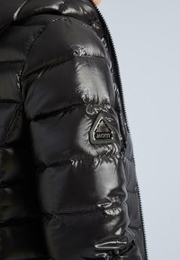 Juodas pūkinis švarkas, pagamintas iš blizgaus medžiagos, su trikampiu prekės ženklu ant kairiojo rankovės ir užtrauktuku.