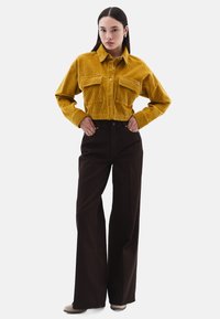 Žlutá kordová zkrácená košile se dvěma předními kapsami, zkombinovaná s vysoce pasovými širokými kalhotami tmavě hnědé barvy. Měkká textura a zapínání na knoflíky.