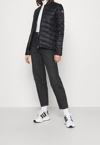 Svart puffjacka med quiltat mönster, dragkedjestängning och logotyp på ärmen. Matchad med mörka jeans och svart-vita sportskor.