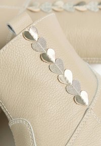 Beige leather boots with a decorative heart-shaped gold and silver accent along the side. Features stitching and a rear pull tab.
