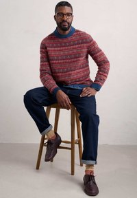 Maglione lavorato a maglia con motivo geometrico multicolore, colletto a coste blu, jeans blu scuro e scarpe bordeaux. Sgabello in legno marrone e sfondo chiaro.