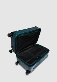 Maleta con ruedas de color verde azulado, con asa retráctil, compartimentos con cremallera y textura suave. El interior cuenta con forro negro y bolsillos organizadores.