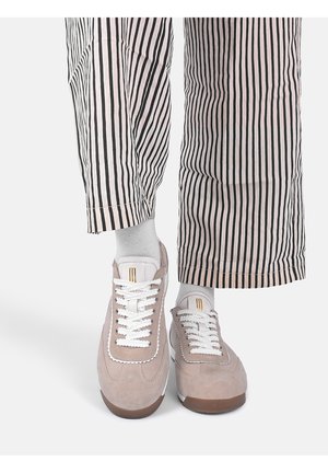 Füße mit weißen Socken, beigen Turnschuhen mit weißen Schnürsenkeln und schwarz-weiß vertikal gestreiften Hosen vor einfachem Hintergrund.