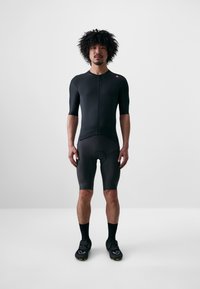 Completo da ciclismo nero con maniche corte, zip frontale e area imbottita per la seduta. Include scarpe nere con accenti verdi e calzini coordinati.