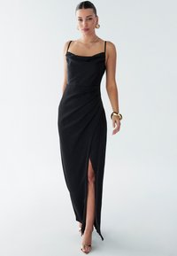 Black satin evening dress with spaghetti straps, cowl neckline, draped detailing, and a high slit. Paired with metallic strappy heels.