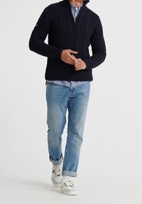 Marinblå kabelstickad tröja med hög krage, i kombination med en rutig skjorta och ljusblå jeans. Vita sneakers med detaljer fulländar outfiten.