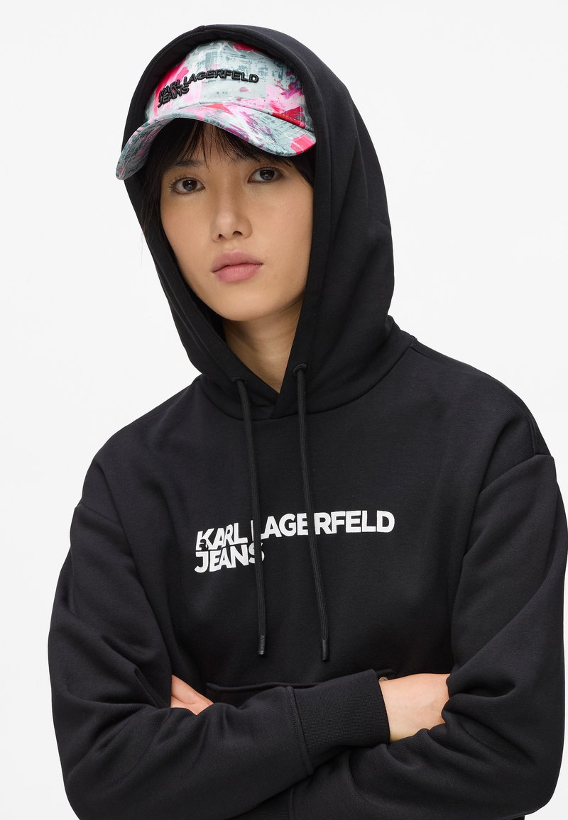 Sudadera negra con un logo blanco, que incluye un cordón. Combinada con una gorra multicolor que muestra gráficos y texto, predominantemente en rosa y gris.