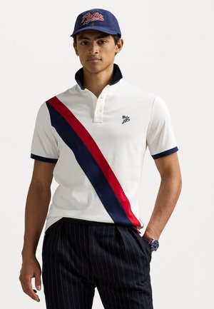 Witte poloshirt met een diagonale rode en marineblauwe streep, voorzien van een gevouwen kraag en een knoopsluiting met twee knopen; gecombineerd met gestreepte broek.
