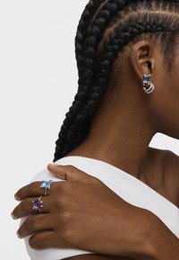 Par de anillos de plata con una gran piedra rectangular azul y una piedra púrpura, llevados en los dedos junto a un pendiente de aro de plata.