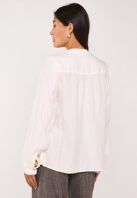 Camicia bianca a maniche lunghe con vestibilità rilassata, dettaglio plissettato sul retro e polsini con bottone. Indossata con pantaloni grigi a fantasia.