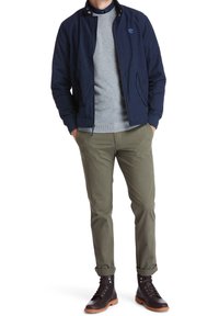 Man wearing navy jacket, grey sweater, olive green pants, and dark brown leather boots standing with hands in pockets.