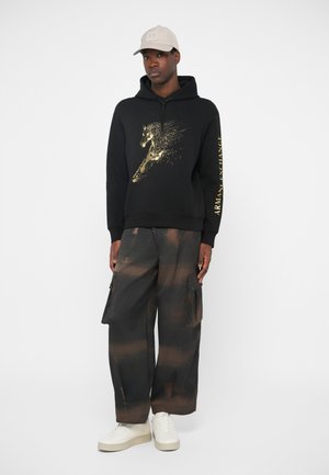 Sort sweatshirt med gyldent hesteprint, parret med løse sorte cargobukser med brune striber og hvide sneakers. Beigefarvet kasket fuldender looket.