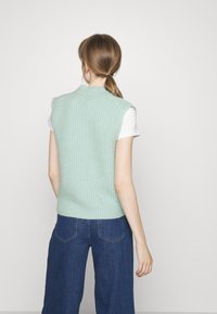 Muntgroene gebreide mouwloze vest met een geribbelde structuur en een platte kraag, gedragen over een witte t-shirt en gecombineerd met donkerblauwe wijde jeans.