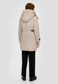 Lichtbeige gewatteerde jas met een grote capuchon, een taillering en zwarte accenten. Heeft lange mouwen en een gladde, gestructureerde stof.