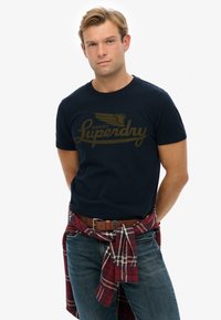 Camiseta de algodón azul marino con un diseño de logo dorado. Combinada con jeans de lavado claro y una camisa de cuadros roja atada a la cintura.