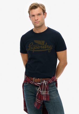 Camiseta de algodón azul marino con un diseño de logo dorado. Combinada con jeans de lavado claro y una camisa de cuadros roja atada a la cintura.