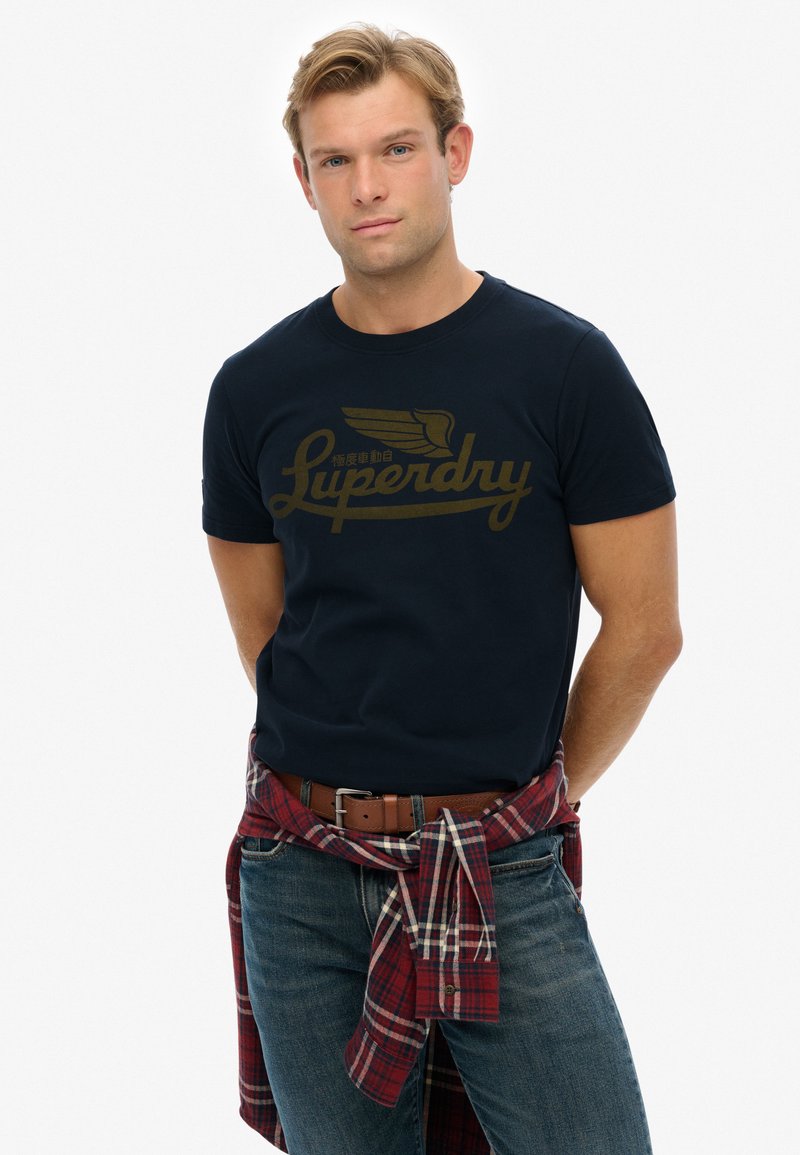 Camiseta de algodón azul marino con un diseño de logo dorado. Combinada con jeans de lavado claro y una camisa de cuadros roja atada a la cintura.