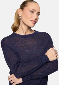 Femme aux cheveux blonds attachés en queue de cheval, portant des boucles d'oreilles argentées en anneau et un pull en tricot bleu foncé, croisant les bras et regardant vers la droite.