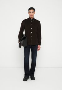 Brun kordfløyelskjorte med knappeåpning, parret med mørkeblå denimjeans og svarte lærstøvler. Modellen holder en svart veske.