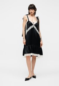 Γυναίκα με σκούρα μαλλιά που φοράει μαύρο σατέν φόρεμα με λευκή δαντέλα στο τελείωμα και μαύρα μυτερά παπούτσια με χαμηλό τακούνι, στέκεται μπροστά σε απλό φόντο.