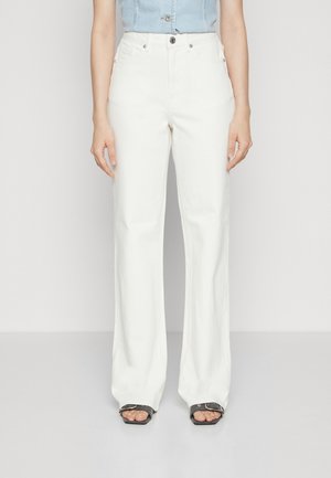 Persona con pantalones blancos de pierna ancha y cintura alta y una camiseta sin mangas azul claro, de pie descalza sobre un fondo claro y sencillo.